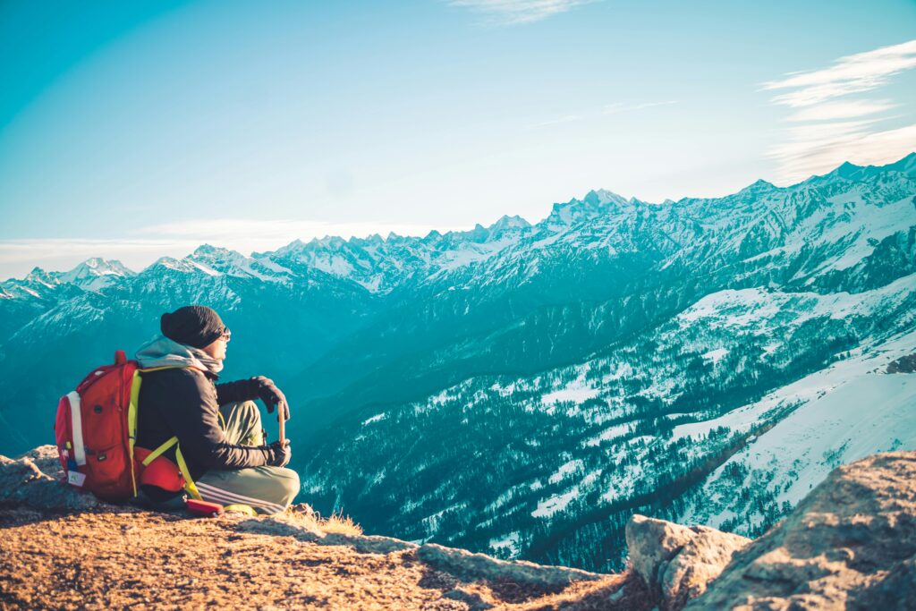A lone backpacker sitting on a mountain summit admiring the snowy peaks of the Himalayas.